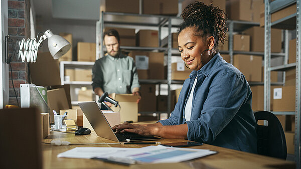 Verschiedene männliche und weibliche Lagerbestandsmanager, die sich unterhalten, Laptops benutzen und den Einzelhandelsbestand überprüfen. Regalreihen voller Kartonverpackungen im Hintergrund. 