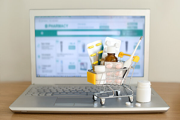 Warenkorb-Spielzeug mit Medikamenten vor dem Laptop-Bildschirm mit Apotheke-Website auf ihm. Tabletten, Blisterpackungen, medizinische Flaschen, Thermometer-Set. Gesundheitsfürsorge und Internet-Shopping. Bilschirm Laptop mit Warenkorb Online Apotheke
