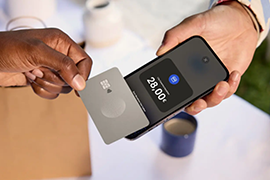 : Eine Person hält eine silberne Kreditkarte auf ein Smartphone. Das Display zeigt einen Einkaufsbetrag